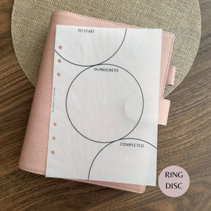 May include: A light pink textured planner with a white insert page featuring three large black circular outlines labeled TO START, IN PROGRESS, and COMPLETED. The paper has a vertical row of six circular punch holes along the left side.