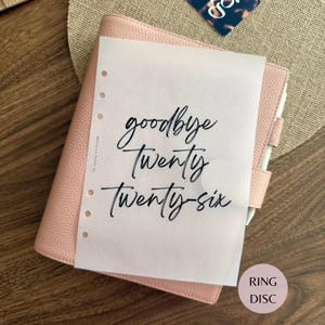 May include: A light pink textured leather planner with a translucent vellum insert featuring the black cursive text goodbye Twenty Twenty-six. The planner includes a side pen loop and a series of circular punch holes along the left edge of the page.