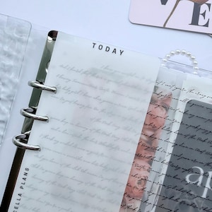 May include: A clear planner insert with the word "TODAY" printed at the top. The insert has handwritten text and is held in a binder with silver rings. The words "MARCELLA PLANS" are printed on the side.