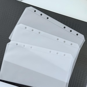 May include: Clear plastic binder pockets with reinforced edges and pre-punched holes for easy insertion into a binder. The pockets are stacked on a black textured surface, with a white background visible.