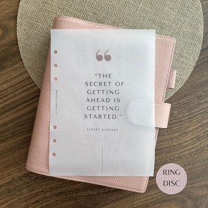 Puede incluir: Una agenda de color rosa claro con textura de cuero, cierre a presión y una hoja de papel vegetal translúcido con la cita: "The secret of getting ahead is getting started." de Elbert Hubbard. La página tiene seis agujeros perforados.