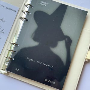 以下が含まれることがあります： クリーム色のプランナーが開き、ハロウィーンをテーマにした白黒のインサートがページにあります。インサートには、魔女の帽子をかぶった人のシルエットと「Happy Halloween!」と「CAMERA! PLAY」という文字が描かれています。タイムスタンプは00:23:59です。
