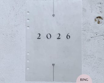 May include: A translucent vellum dashboard for a ring-bound planner featuring the text 2026 in a black serif font. The design includes a vertical line with small heart icons above and below the year, and six circular punch holes on the left side.