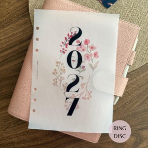 Puede incluir: Una hoja de papel vegetal translúcido para agenda con el año 2027 en grandes números negros con serifa, rodeada de delicadas ilustraciones en acuarela de flores silvestres rosas y beige, con seis agujeros perforados y el texto Marcella Paper Co.