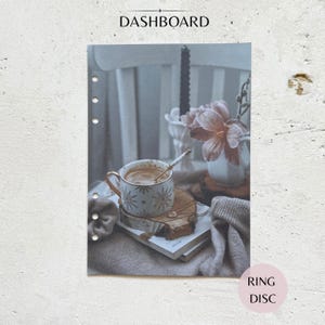 Può includere: Un dashboard per agenda rettangolare con sei fori punzonati lungo il bordo sinistro. L'immagine mostra una tazza bianca con motivi a stella dorati, un sottobicchiere in legno, un libro, un vaso bianco con un fiore rosa e una candela nera.