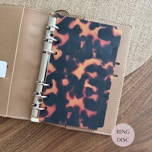 May include: A beige textured planner with a six-ring silver metal binder mechanism. Inside, a dark brown and orange tortoiseshell patterned plastic divider is attached to the rings. The planner features a smooth interior finish and slots.
