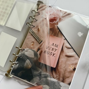 May include: A clear binder with gold-colored rings, holding a pink notebook that reads "I AM VERY BUSY." The binder also contains a photo with a teddy bear, a laptop, and a rose. The binder is held by a person.