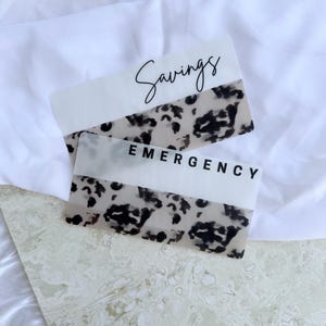 Puede incluir: Dos tarjetas de presupuesto rectangulares de acrílico. La tarjeta superior dice "Savings" en cursiva. La tarjeta inferior dice "Emergency" en sans-serif. Ambas tarjetas tienen una sección superior blanca y una sección estampada con manchas negras y beige.