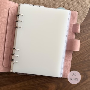 May include: A light pink A5 ring binder with a textured leather-effect cover, featuring a six-ring silver metal mechanism. Inside, a translucent white divider page is visible, along with monthly tabbed dividers labelled Jan through Dec.