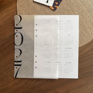 May include: A white paper planner insert for the year 2027 featuring a translucent vellum overlay. The left side displays the year 2027 in large black font, while the right side lists all twelve months with their corresponding dates.