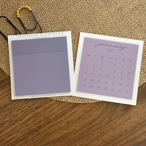 May include: Two square transparent sticky note pads in light purple. The left pad is labeled Transparent Sticky Notes Purple Rain Marcella Paper Co. The right pad features a January 2027 calendar grid with days of the week printed in dark gray.