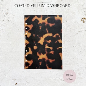 May include: Coated vellum dashboard with a tortoiseshell pattern in shades of brown and amber. The planner insert has pre-punched holes for ring binders. The text "COATED VELLUM DASHBOARD" is at the top.