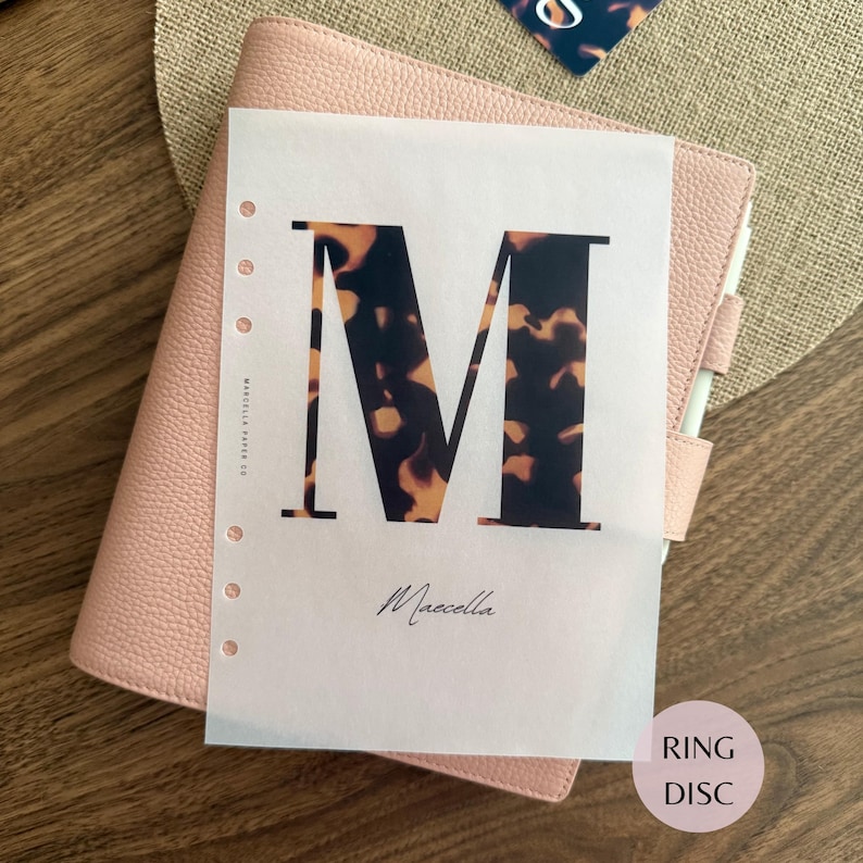May include: A translucent planner insert page featuring a large letter M with a brown and black tortoiseshell pattern. The text Marcella Paper Co is printed vertically on the left side, and the name Marcella is printed in script below the letter M.
