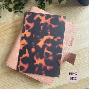 Può includere: Un foglio rettangolare con un motivo tartarugato marrone scuro e arancione posizionato sopra un raccoglitore ad anelli rosa chiaro testurizzato. Il raccoglitore presenta una superficie granulata, una linguetta di chiusura e anelli metallici.