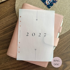May include: A light pink leather-textured planner with a translucent vellum sheet overlay. The vellum features the year 2027 in black text, small heart icons, and vertical lines. The planner has a side tab closure and a series of punched ring holes.