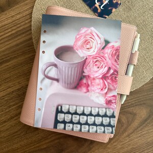 Puede incluir: Una agenda de cuero rosa claro con un bol&iacute;grafo blanco y una inserci&oacute;n decorativa que muestra una taza rosa claro, un ramo de rosas rosas y un teclado de m&aacute;quina de escribir. La agenda tiene un acabado texturizado y una trabilla para bol&iacute;grafo.