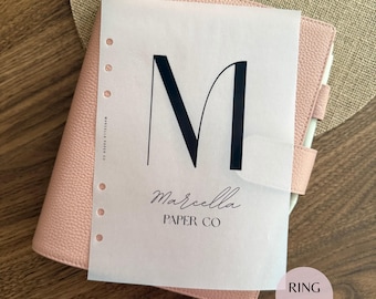 May include: A light pink textured leather ring binder planner featuring a translucent vellum sheet with a large black letter M, the text Marcella, and the words PAPER CO printed in black. The sheet has a vertical row of circular punch holes.