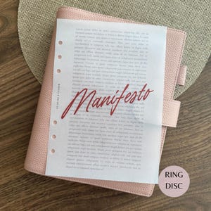 May include: A light pink textured leather planner with a strap closure. A white sheet of paper with punched holes on the left side is placed on top. The paper features printed text and the word Manifesto in large red script. A circular tag reads RING DISC.