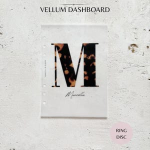 May include: A translucent vellum dashboard for a ring-bound planner featuring a large letter M with a brown and black tortoiseshell pattern. The text Marcella is printed in a black script font below the letter, with small punch holes on the left side.