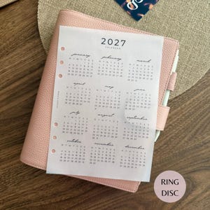 May include: A light pink leather-textured planner with a white 2027 calendar insert featuring twelve months of dates in a grid format. The planner has a side pen loop and a series of circular holes along the left edge for ring binding.