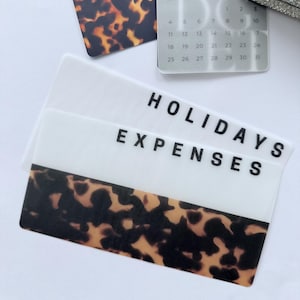 May include: Clear plastic labels with the words "HOLIDAYS" and "EXPENSES" in black. The labels feature a tortoiseshell pattern. A small calendar for January 2026 is also visible, along with a tortoiseshell patterned card.