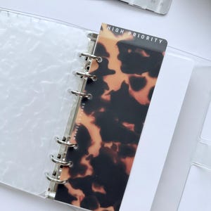 May include: A clear, plastic binder with silver rings holds a patterned card with the text "HIGH PRIORITY." The card has a tortoiseshell design. A calendar card is visible in the background, along with a matching tortoiseshell card.