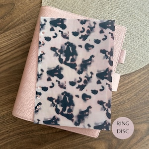 May include: A translucent planner dashboard featuring a dark brown and black tortoiseshell pattern on a light beige background. The rectangular sheet has a vertical row of circular punch holes along the left edge and is placed over a light pink binder.