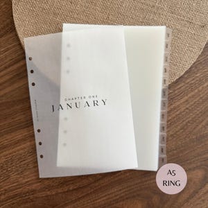 May include: A set of translucent A5 planner inserts featuring a frosted divider page with the black printed text CHAPTER ONE JANUARY and a side tabbed index with the months of the year from January to December printed in black on each tab.