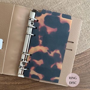 May include: A beige leather-textured planner binder featuring a silver six-ring metal mechanism and a translucent divider with a dark brown and amber tortoiseshell pattern. The planner includes a small circular label with the text RING DISC.