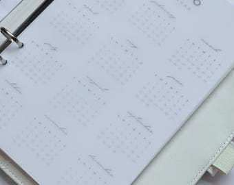 May include: A white planner open to a 2026 calendar page. The calendar is printed in light gray with the year at the top and months labeled in a cursive font. The planner has silver rings and a white leather cover.