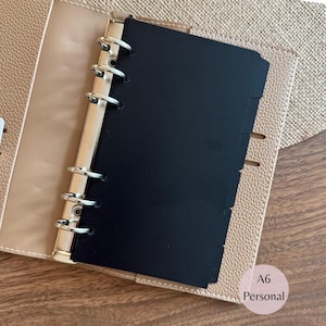 May include: A beige textured leather personal planner with a six-ring silver metal binder mechanism. The planner contains a solid black divider page with tabbed edges. The text A6 Personal is printed in the bottom right corner of the image.