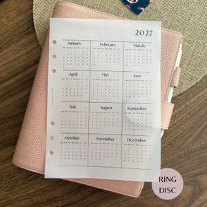 May include: A light pink textured leather planner with a white paper insert featuring a 2027 calendar year overview. The calendar page displays all twelve months from January to December in a grid format with punched holes along the left side.