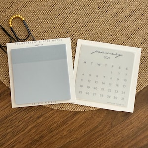 May include: Two square transparent sticky note pads on white backing cards. One pad is solid light blue labelled 'TRANSPARENT STICKY NOTES HEATHER'. The other is a translucent January 2027 calendar with grey text and grid lines, both marked 'MARCELLA PAPER CO'.