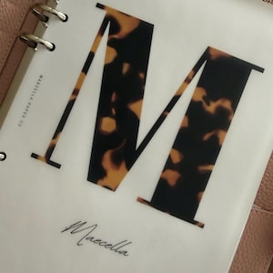 May include: A translucent planner divider featuring a large letter M with a brown and black tortoiseshell pattern. The text Marcella Paper Co is printed vertically on the left, and the word Maecella is written in a cursive script at the bottom.
