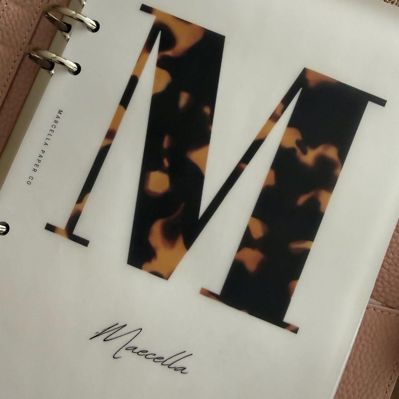 May include: A translucent planner divider featuring a large letter M with a brown and black tortoiseshell pattern. The text Marcella Paper Co is printed vertically on the left, and the word Maecella is written in a cursive script at the bottom.
