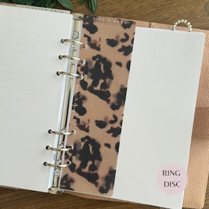 May include: A translucent planner dashboard featuring a brown and black tortoiseshell pattern. The top edge displays the text LIKE A BOSS, and the side includes the text MARCELLA PAPER CO. It is secured within a six-ring metal binder mechanism.