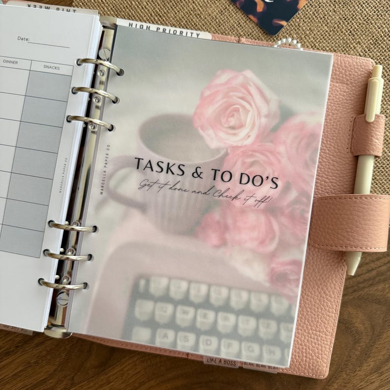 Puede incluir: Una carpeta de cuero rosa empolvado con un mecanismo met&aacute;lico de seis anillas que contiene una hoja de planificaci&oacute;n. La hoja presenta un gr&aacute;fico floral con una taza de caf&eacute; y una m&aacute;quina de escribir, con el texto TASKS & TO DO'S Get it done and Check it off.