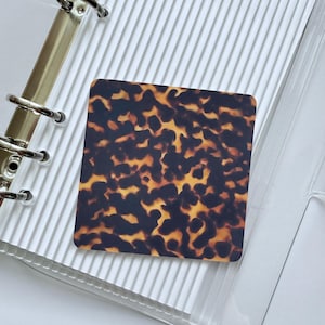 May include: A square, tortoiseshell-patterned card, featuring a mix of brown and amber tones, rests on a clear, ridged surface. The card's design resembles the shell of a tortoise. The background includes a binder with silver rings.