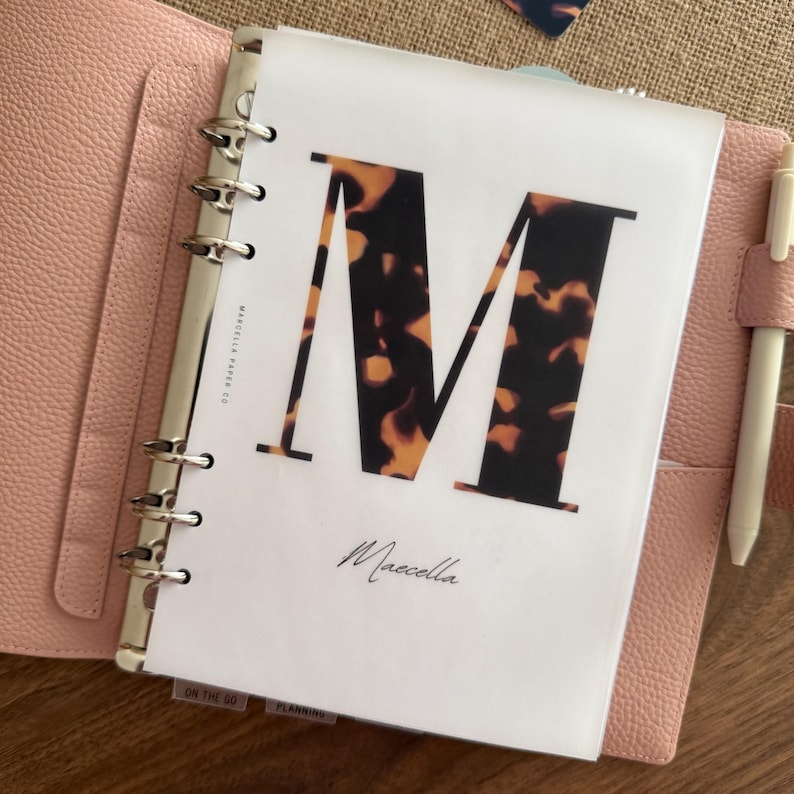 May include: A light pink leather binder with a silver six-ring mechanism containing a white page featuring a large letter M in a brown and black tortoiseshell pattern. The text Marcella Paper Co and the name Marcella are printed on the page.