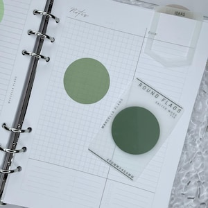 Può includere: Un'agenda con carta a quadretti, un adesivo rotondo verde e una linguetta trasparente con la scritta "IDEAS". È visibile anche una confezione di bandierine rotonde, 4,5 cm x 4,5 cm. L'agenda è aperta su una pagina con la parola "Note".