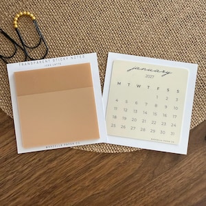 May include: Two square sheets of transparent sticky notes on white backing cards. One sheet is solid tan labeled Iced Latte, and the other is a cream-colored January 2027 calendar. Both feature the text Marcella Paper Co at the bottom.