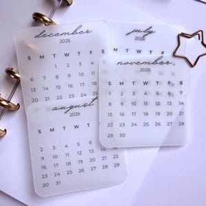 Op de afbeelding: Transparante acryl kalenderinzetstukken voor 2026, met de maanden december, augustus, juli en november. De maanden zijn in cursief schrift geschreven, met een stervormige paperclip. Gouden binderringen houden de kalenderpagina's bij elkaar.
