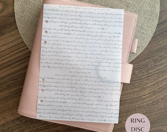 Puede incluir: Una agenda de textura de cuero rosa claro con cierre de correa lateral. Sobre ella descansa una hoja rectangular de papel blanco con líneas repetidas de texto negro en cursiva y una columna vertical de seis agujeros perforados a la izquierda.