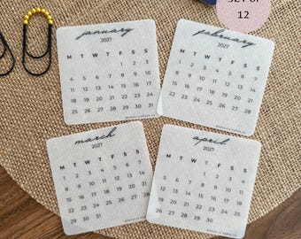 May include: A set of 12 translucent square calendar cards for the year 2027, featuring the months of January, February, March, and April with black text and grid layouts. The cards include the text SET of 12 and MARCELLA PAPER CO.