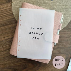 May include: A light pink textured leather planner with a white translucent vellum sheet featuring the black text IN MY DELULU ERA. The planner includes a white pen and a circular tag with the text RING DISC. The planner has a side strap closure.