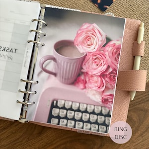 Puede incluir: Una agenda de anillas de cuero rosa claro con un bol&iacute;grafo crema en un lazo lateral. La agenda abierta muestra una p&aacute;gina de tareas y una foto con una taza rosa claro, una m&aacute;quina de escribir rosa y un ramo de rosas rosas sobre fondo blanco.