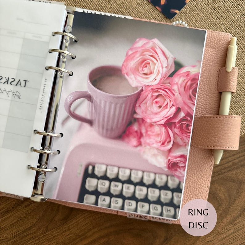 Puede incluir: Una agenda de anillas de cuero rosa claro con un bol&iacute;grafo crema en un lazo lateral. La agenda abierta muestra una p&aacute;gina de tareas y una foto con una taza rosa claro, una m&aacute;quina de escribir rosa y un ramo de rosas rosas sobre fondo blanco.