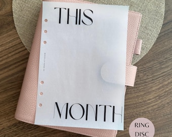 May include: A light pink textured leather planner with a translucent vellum dashboard insert. The insert features the black printed text THIS MONTH and a vertical row of six punched holes along the left side, with small text reading MARCELLE PAPER CO.