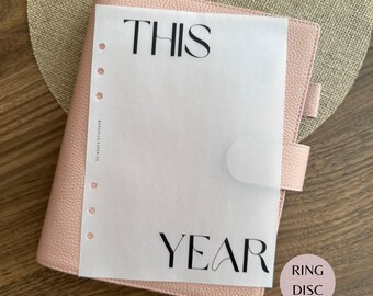 May include: A light pink textured leather planner with a translucent vellum sheet overlay. The sheet features the black text THIS at the top and YEAR at the bottom, with a vertical row of six circular punch holes along the left side edge.