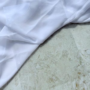 Könnte beinhalten: Nahaufnahme eines weißen, durchscheinenden Stoffes mit zarter Textur. Der Stoff ist drapiert und erzeugt weiche Falten und Schatten. Der Hintergrund ist eine hellfarbige Oberfläche mit einem dezenten Muster.